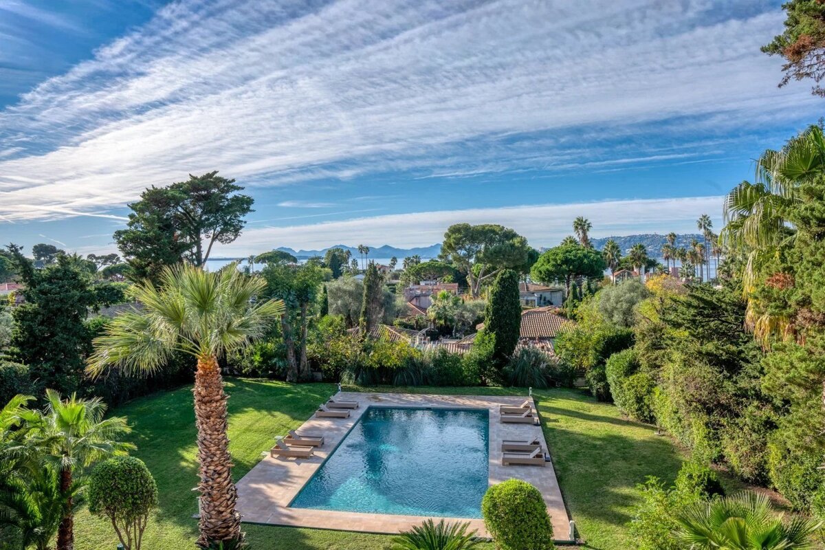 Aerial view of a private pool in a lush garden with palm trees, overlooking the sea and distant mountains under a blue, cloudy sky.