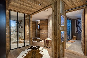 Rustic wooden chalet interior with a glass wine cellar, cowhide rugs, ski posters, and a spa area with a large portrait.