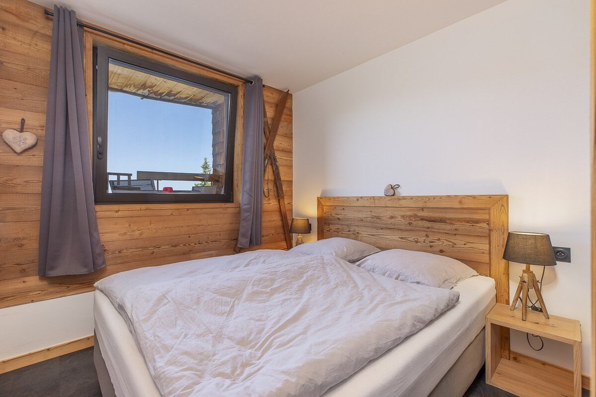 A cozy bedroom with wood-paneled walls, a large window, a bed with light grey bedding, and ski decor, evoking a warm, rustic cabin feel.