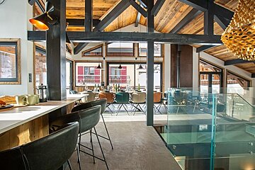 Modern alpine chalet interior with rustic wooden beams, a breakfast bar, glass railing, and dining area with snowy views.