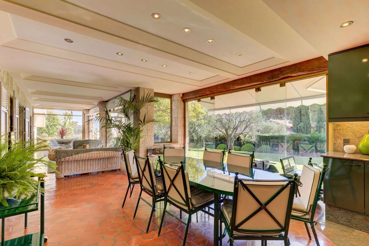 Bright room with terracotta hex tile floors, a dining table, and a living area, all opening to a lush garden through expansive glass windows.