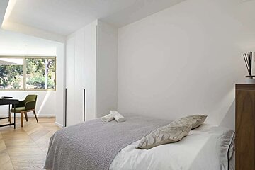 A bright, minimalist bedroom with a bed, white walls, wooden floor, and a window overlooking green trees. A simple desk setup is in the background.