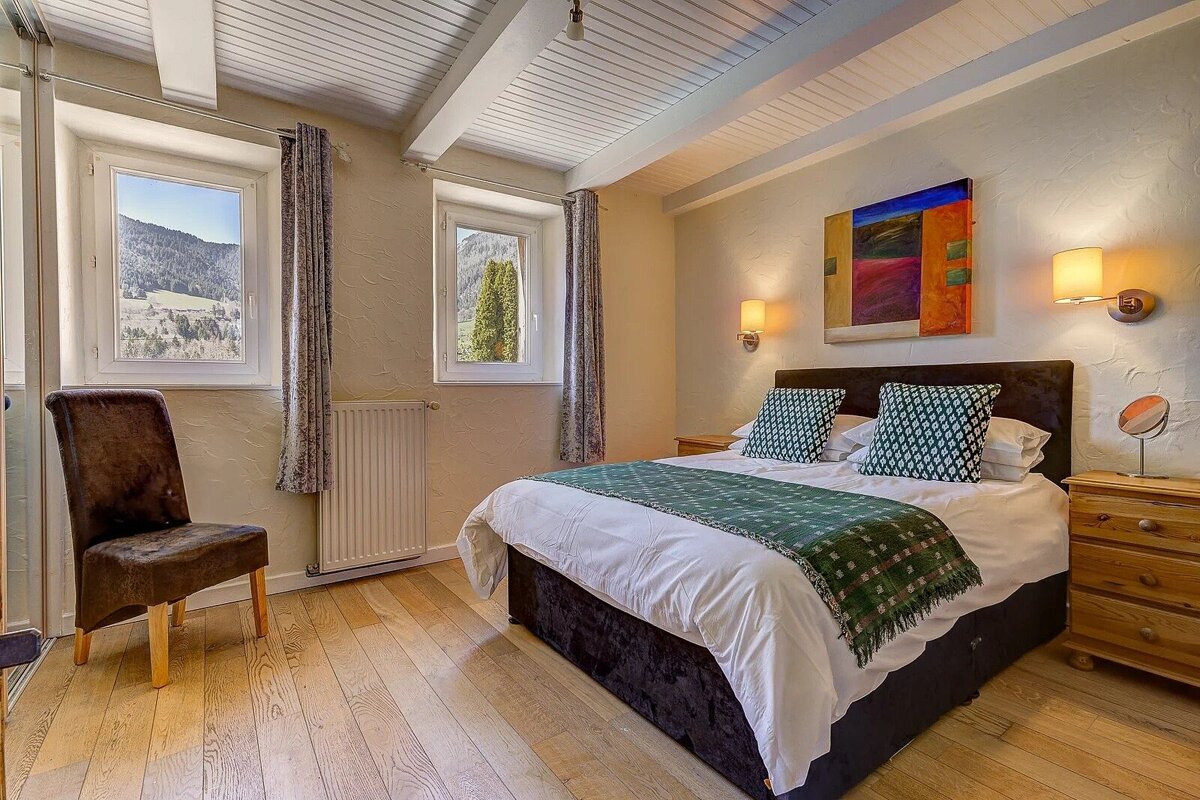 A cozy bedroom features a double bed, wooden floors, exposed beams, and two windows with scenic mountain views. Wall art and lamps adorn the space.