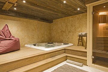 A relaxing spa room with a hot tub, sauna, and bean bag chair. Features rustic wooden ceiling, steps, and textured walls, creating a cozy ambiance.