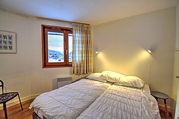A bright bedroom with two single beds, a window overlooking a snowy mountain village, and warm wall lights creating a cozy atmosphere.