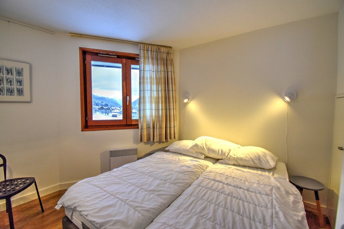 A cozy bedroom featuring twin beds pushed together, warm wall lights, and a window overlooking a snowy mountain landscape.