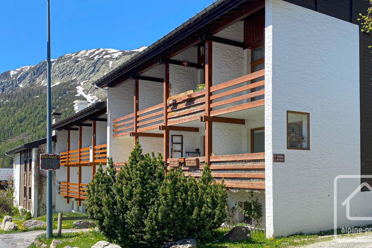 A white chalet-style building with wooden balconies, green shrubs, and snow-capped mountains under a clear blue sky.
