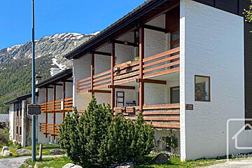 A white building with wooden balconies stands before a snowy mountain range. Green shrubs are in the foreground under a clear blue sky.