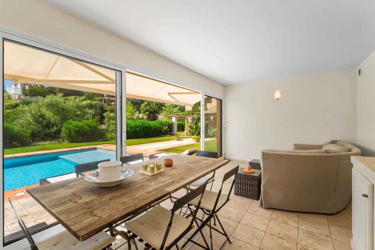 Indoor dining room with a rustic wooden table opens through glass doors to a patio with a swimming pool and lush garden, shaded by awnings.