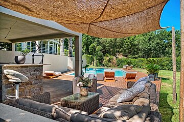 A sunny outdoor patio features a swimming pool, woven lounge furniture, a bar, and a natural straw shade canopy, surrounded by lush greenery.