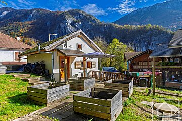 A small house with wooden shutters and planters sits in a sunny, rural mountain setting, featuring clear blue skies and autumn foliage.