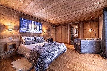 A warm, wooden chalet bedroom featuring a big bed, winter forest artwork, plush seating, and an ensuite bathroom.