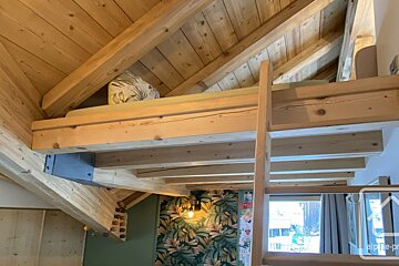 A rustic wooden loft bed with a ladder, featuring exposed beams and an angled ceiling, above a room with tropical leaf wallpaper.