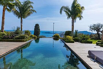 An infinity pool with a wooden deck, flanked by lush gardens and palm trees, offers panoramic views of the blue ocean and distant islands.