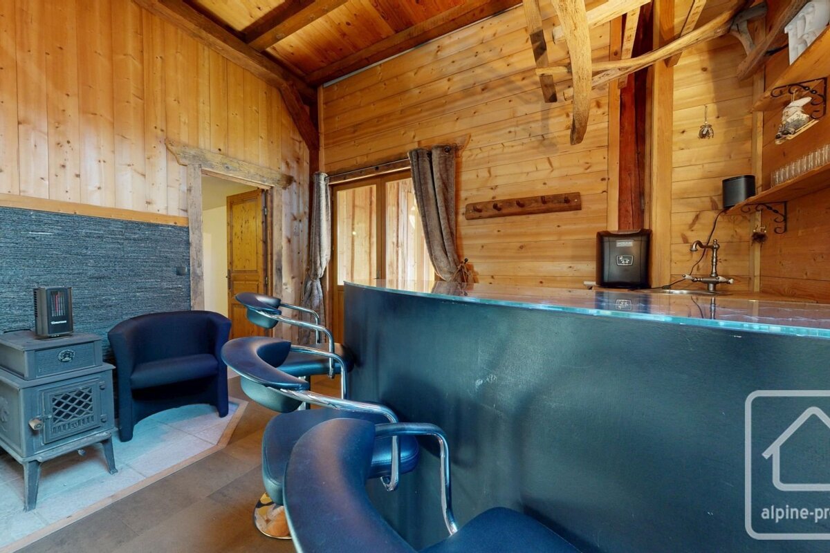 Rustic wood-paneled bar area with a dark counter, blue bar stools, a small black stove, and an armchair, evoking a cozy chalet feel.
