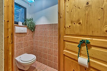 A small, rustic bathroom features brown tiled walls, a white wall-mounted toilet, and a knotty pine door with hanging toilet paper. A plant and artwork add detail.