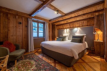 Cozy cabin bedroom with wood-paneled walls, a large bed with white linens, a green armchair, and warm, inviting lighting.