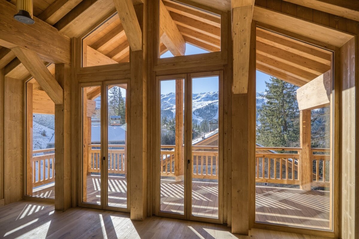 A rustic wooden room with large glass doors and windows opens to a sunlit balcony, revealing a breathtaking view of snowy mountains and pine trees.
