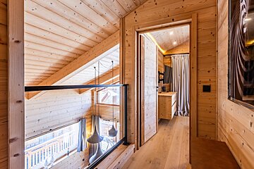 Interior of a modern wooden chalet, featuring a mezzanine with a glass railing overlooking the lower floor and a hallway leading to a bedroom.