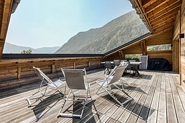A wooden deck with chairs overlooks mountains under a blue sky.
