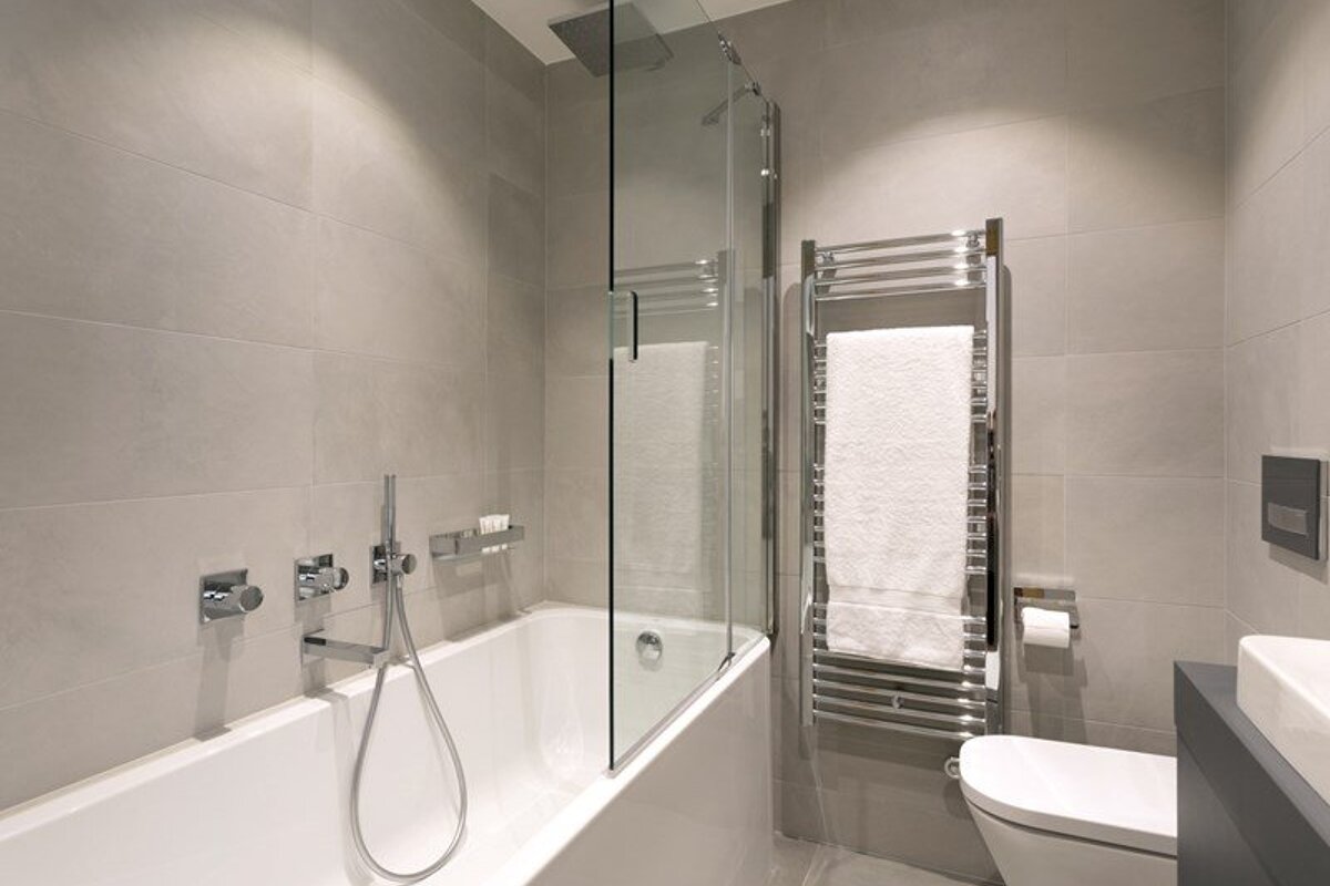 A modern bathroom with a white bathtub, a glass shower partition, a chrome heated towel rack, and light gray tiled walls.