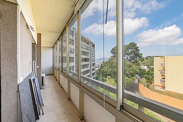 A long, glass-enclosed balcony offers panoramic views of city buildings, lush green trees, and a blue sky. An AC unit is visible inside.