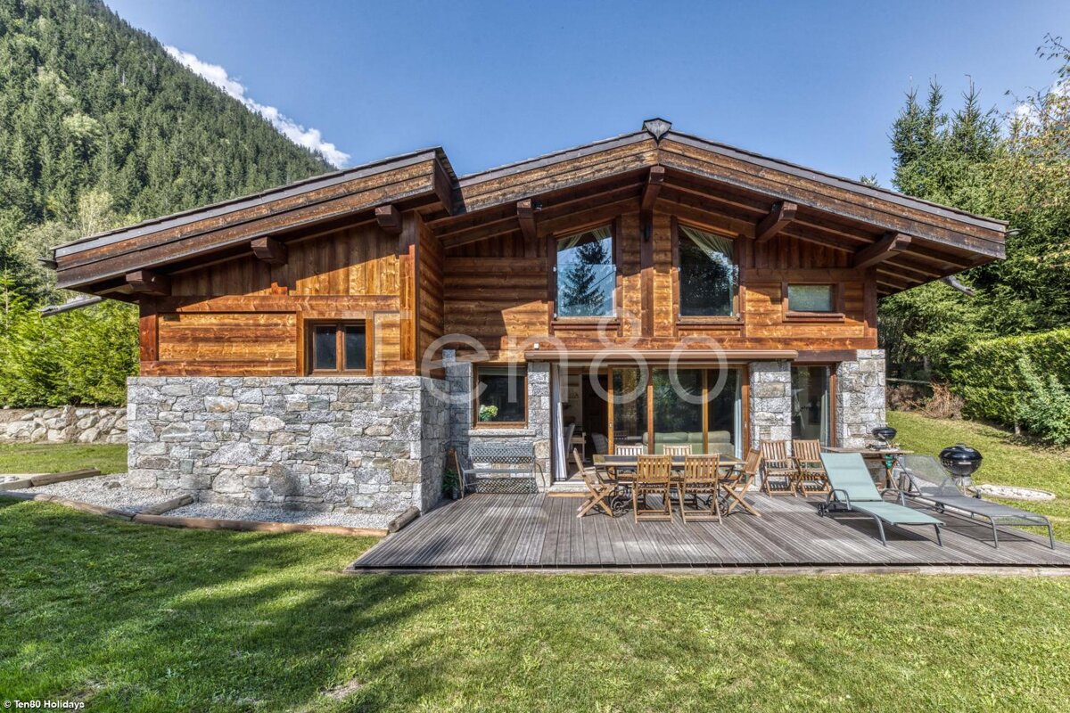 A rustic wooden and stone chalet with a deck, patio furniture, and green lawn, nestled against a forested mountain under a blue sky.