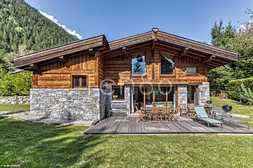 A rustic wooden and stone chalet with a deck, patio furniture, and green lawn, nestled against a forested mountain under a blue sky.