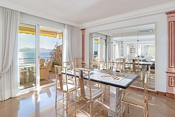 A bright, elegant dining room with gold chairs and a marble table, featuring a stunning balcony overlooking a sunny sea view.