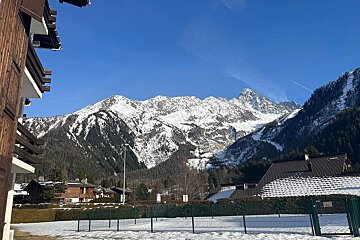 Snow-capped mountains tower over wooden alpine chalets under a bright blue sky.