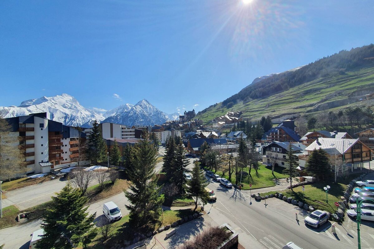 A sunny mountain village scene with snow-capped peaks, green hills, buildings, trees, and roads, under a clear blue sky.