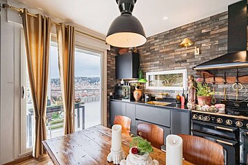 A stylish kitchen and dining area features a rustic brick wall, dark cabinets, a wooden table, and a balcony with expansive city views.