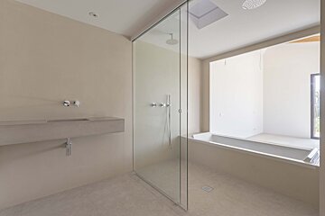 A modern, minimalist bathroom with a glass shower, concrete sink, and built-in bathtub. Neutral tones, skylight, and large window.