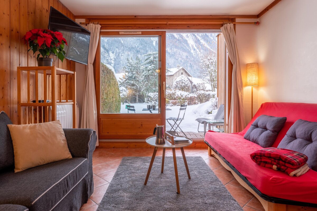 A cozy living room with large glass doors opens to a snowy winter landscape and mountains. Features red and gray sofas, wood paneling, and a poinsettia.