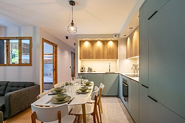 A modern, open-plan kitchen and dining area with light blue-green cabinets, wooden accents, and a set dining table next to a living space.