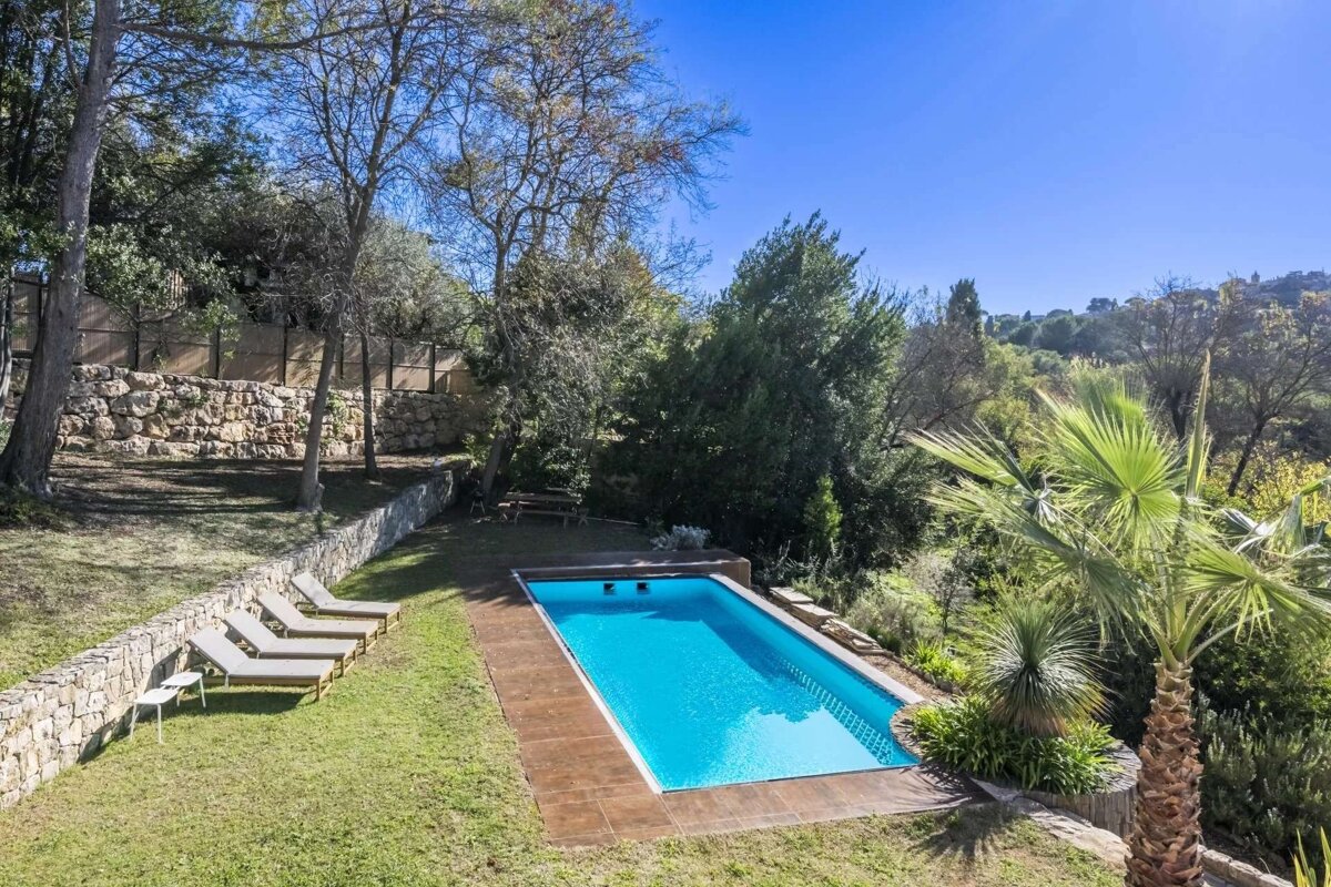 A bright aerial view of a rectangular swimming pool with sun loungers on a grassy terrace, surrounded by lush trees and stone walls under a clear blue sky.