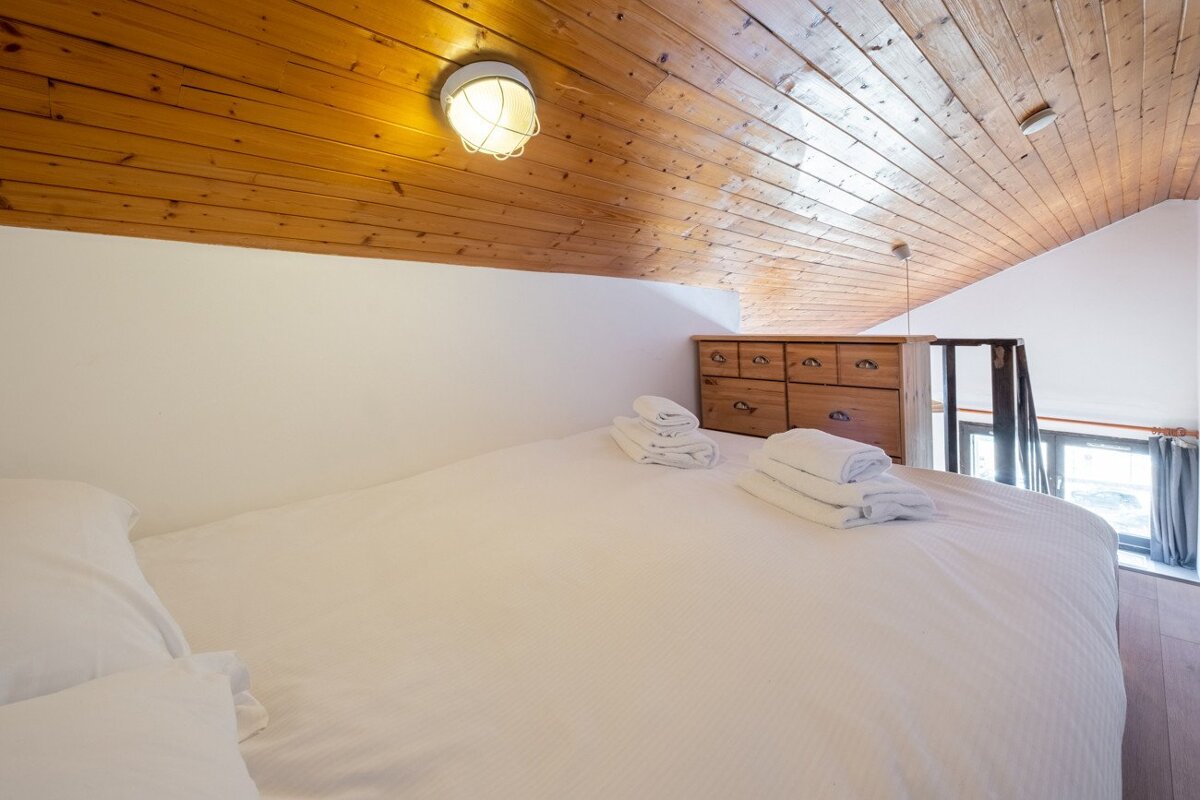 A cozy loft bedroom with a large white bed, wooden sloped ceiling, and a wooden chest of drawers. Fresh towels are neatly stacked on the bed.