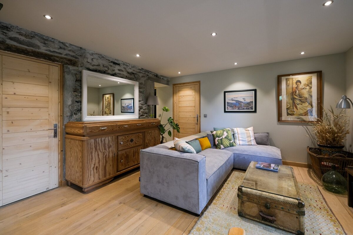 A rustic-modern living room with a grey sectional sofa, exposed stone wall, wooden sideboard, light wood floor, and eclectic art pieces.