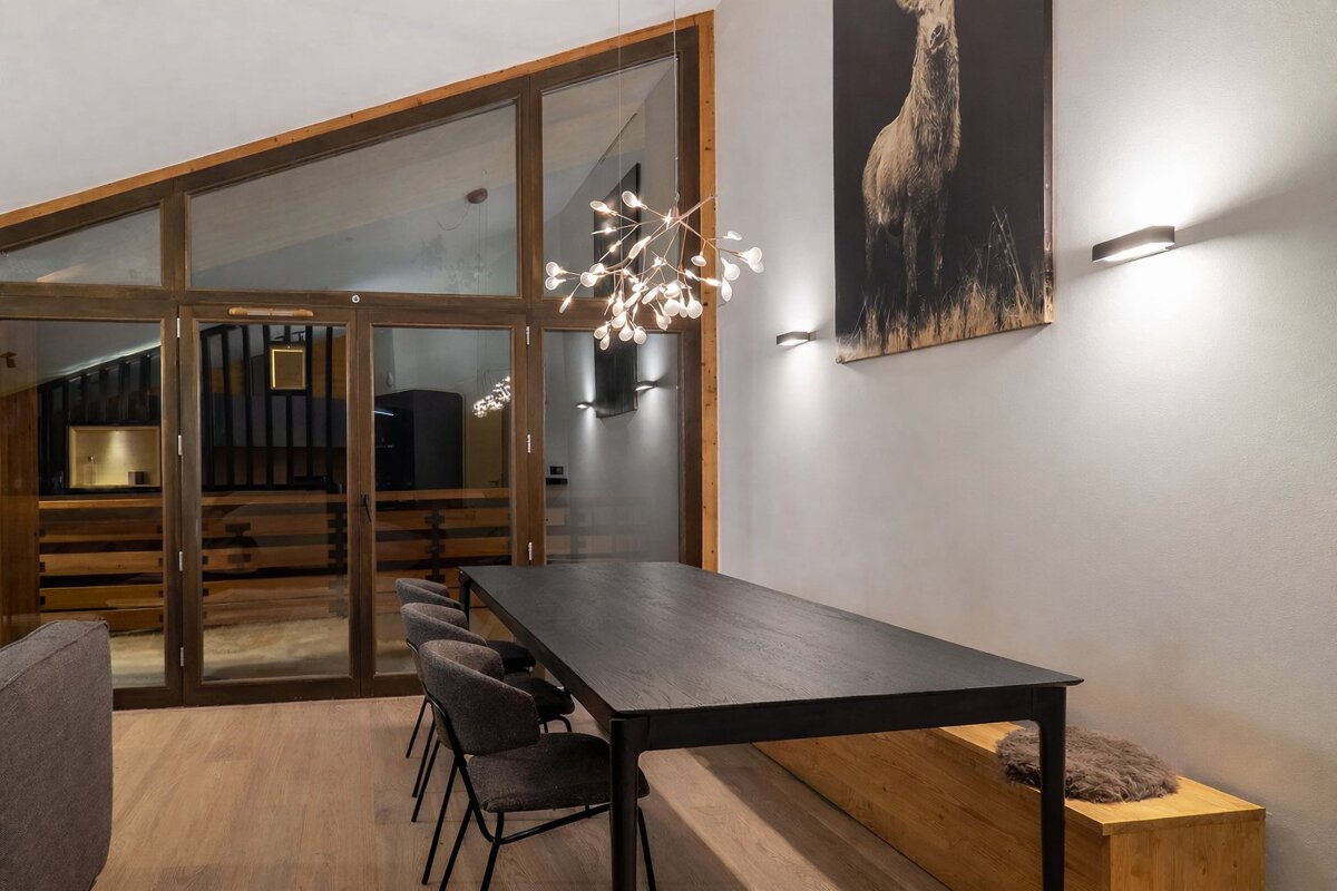 A modern dining area features a long black table, gray chairs, and a branch-like chandelier. Large glass doors and a deer art print adorn the wall.