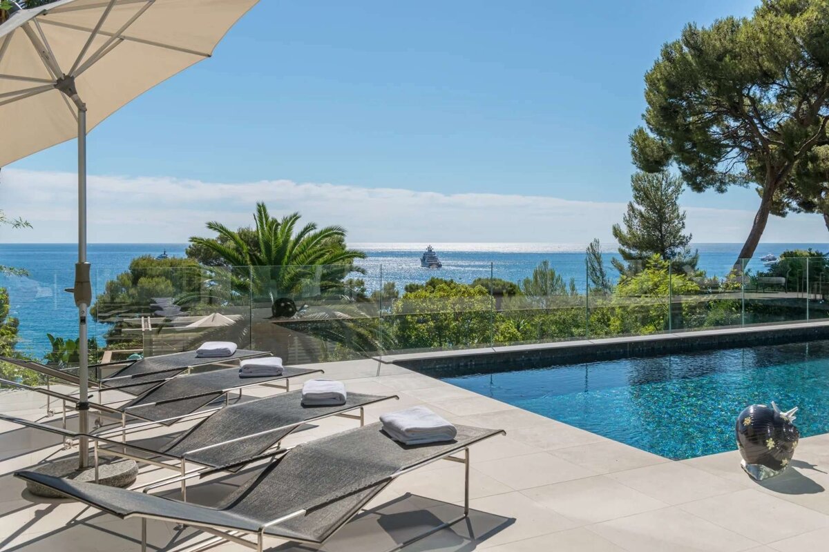 Sunny terrace with lounge chairs and a pool, offering a stunning view of the blue ocean with a yacht, under a clear sky.