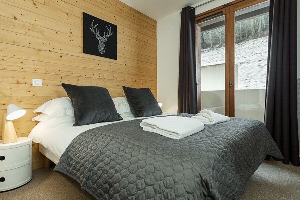 Cozy cabin bedroom with wood-paneled walls, a grey quilted bed, deer art, and glass doors opening to a snowy view.