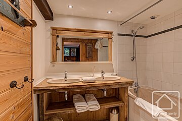 A cozy, rustic bathroom features a wooden double-sink vanity, mirror, and a tiled bathtub/shower. Wood paneling and white tiles create a warm, cabin feel.