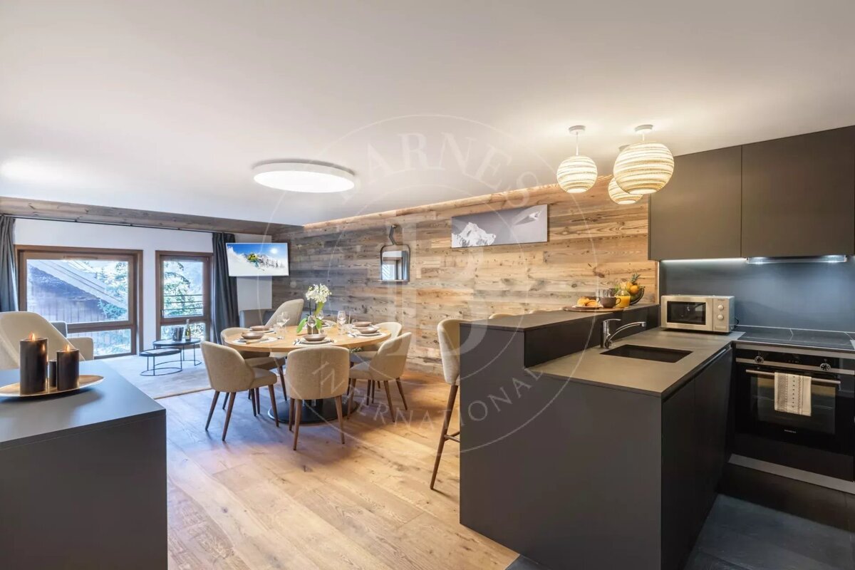 Modern open-plan living space featuring a dark kitchen, wooden accent wall, round dining table, and a cozy living area with large windows.