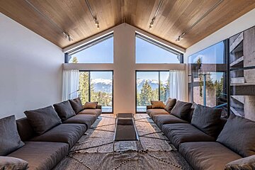 A modern living room with a pitched wooden ceiling and expansive windows showcasing snowy mountains and evergreen forests. Two long couches face each other.