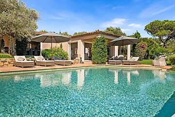 A luxurious turquoise swimming pool with sun loungers and umbrellas, set against a Mediterranean villa and lush greenery under a clear blue sky.