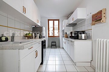 A pristine white galley kitchen featuring white cabinets, tiled floor & backsplash, marble counters, and a wooden window at the end. Modern appliances.