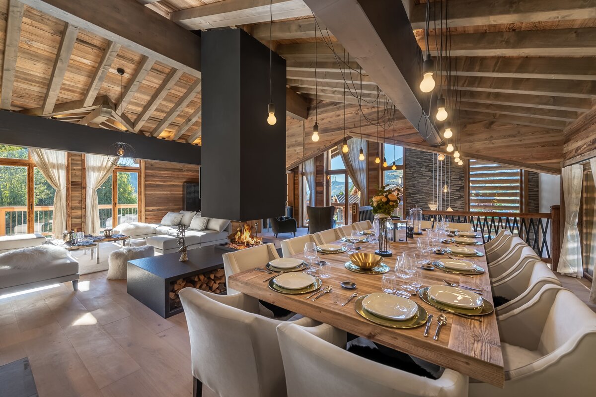 A luxurious chalet interior with exposed wood, a modern fireplace, and a grand dining table set for guests, offering mountain views.