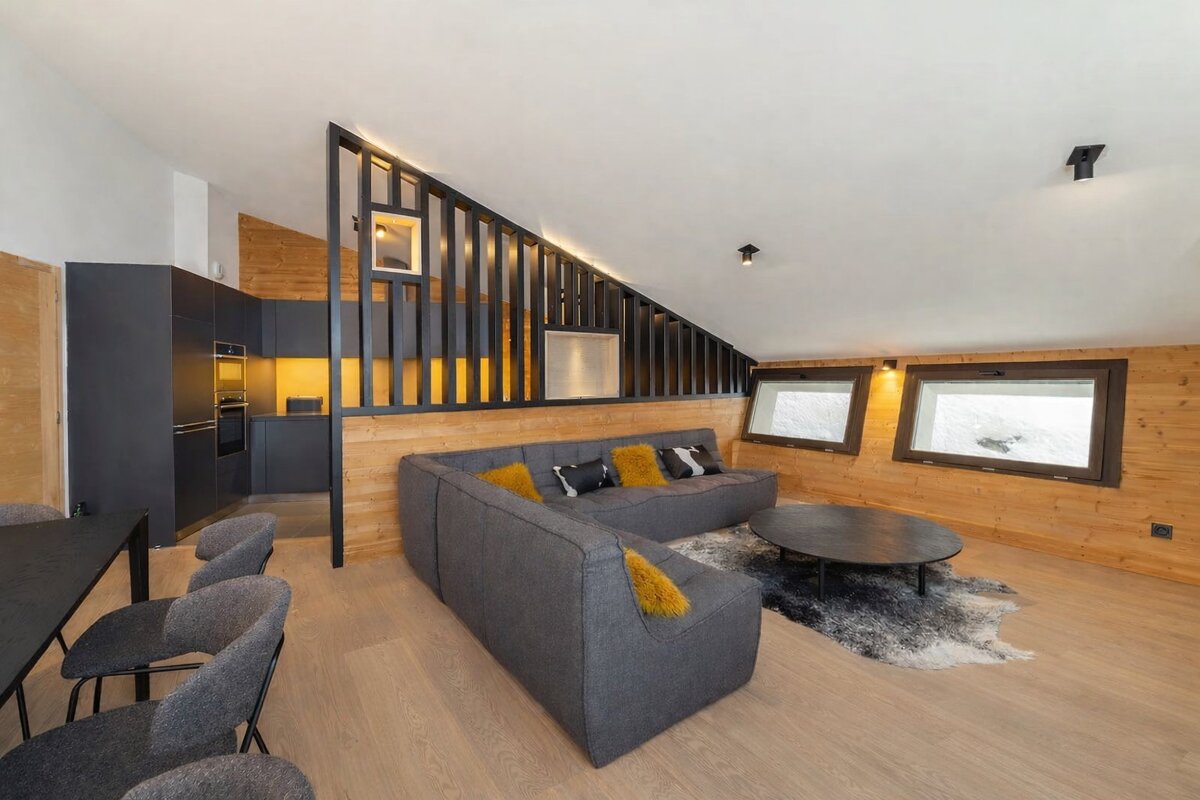 Chic chalet interior: open-plan living room with grey sofa, dark kitchen, wood walls, and snowy window views.