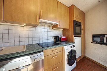 A compact kitchen with light wood cabinets, white tiled backsplash, green countertop, and various appliances including a dishwasher, washing machine, oven, and microwave.