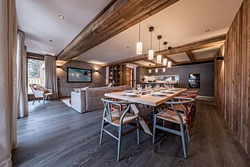 A modern open-plan chalet living space with rustic wooden beams, a large dining table, comfy lounge with TV, and a sleek, gray kitchen.